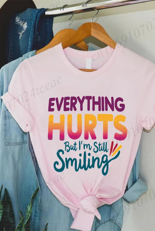 Pink short-sleeve cotton T-shirt with Everything Hurts But I'm Still Smiling quote in colorful bold font, tied at the waist