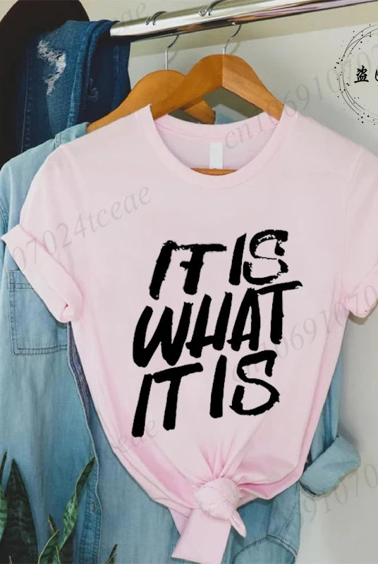 Pink women's short sleeve T-shirt with IT IS WHAT IT IS sarcastic quote graphic, tied at the front, displayed on a hanger.