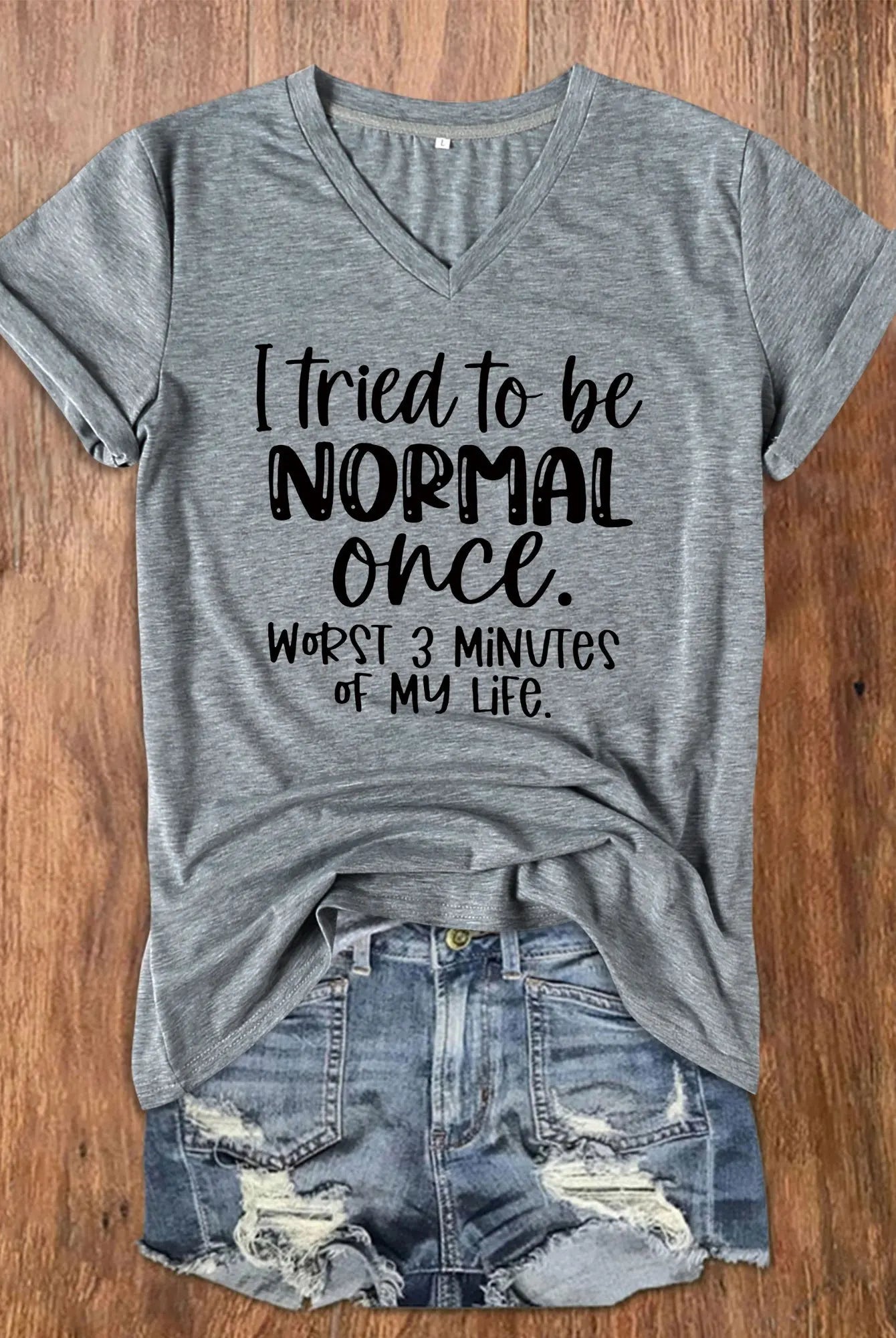 Gray V-neck women's T-shirt with I Tried To Be Normal Once humorous graphic, paired with distressed denim shorts on wooden