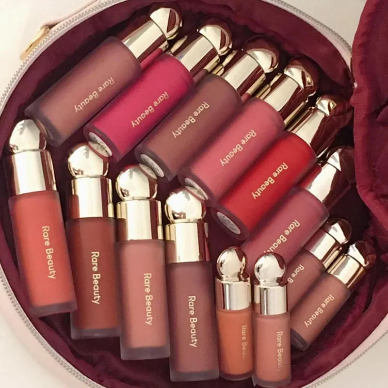 Rare Beauty Soft Pinch Liquid Blush collection in shades of pink, peach, and mauve with gold caps, inside a burgundy fabric-lined bag.